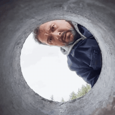 A Man In A Blue Jacket Looks Through A Hole In A Pipe GIF