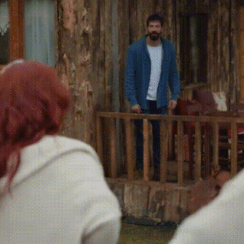 A Man In A Blue Jacket Stands In Front Of A Wooden House GIF