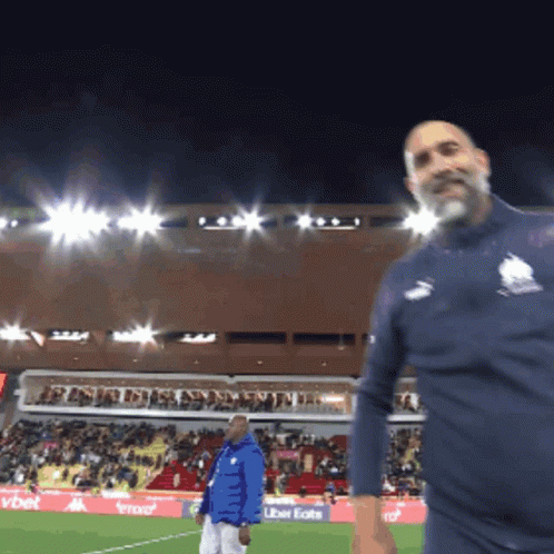 A Man In A Blue Jacket Stands On A Soccer Field In Front Of An Uber Eats Advertisement GIF