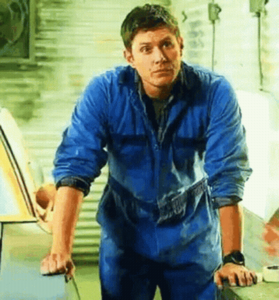 A Man In A Blue Jumpsuit Is Standing In Front Of A Car . GIF
