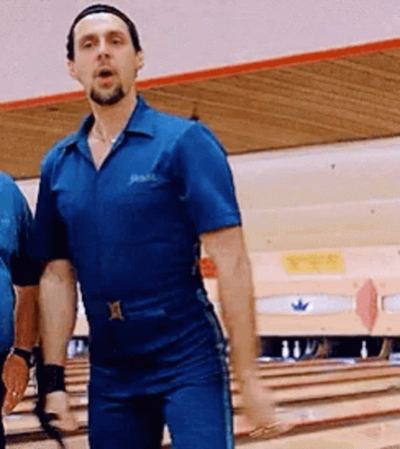 A Man In A Blue Jumpsuit Is Standing On A Bowling Alley With His Hands In The Air . GIF