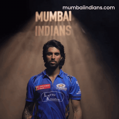 A Man In A Blue Mumbai Indians Jersey Stands In Front Of A Sign That Says Mumbaiindians.com GIF