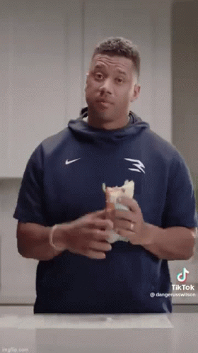 A Man In A Blue Nike Shirt Is Holding A Sandwich In His Hands . GIF