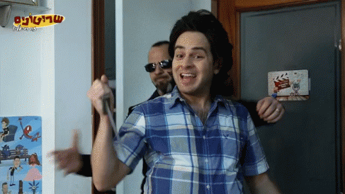 A Man In A Blue Plaid Shirt Is Holding A Microphone In Front Of A Sign That Says ' סרט ' On It GIF