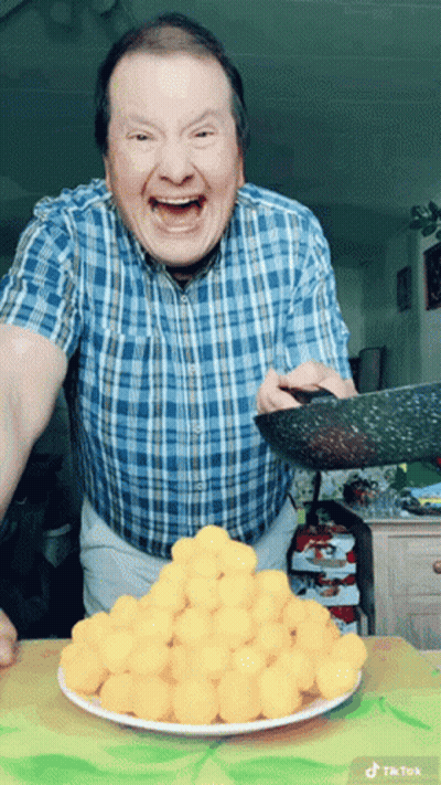 A Man In A Blue Plaid Shirt Is Holding A Pan Over A Plate Of Cheese Balls GIF