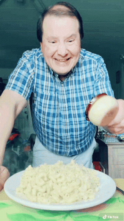 A Man In A Blue Plaid Shirt Is Pouring A Slice Of Onion On A Plate Of Macaroni And Cheese GIF