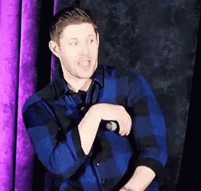 A Man In A Blue Plaid Shirt Is Standing In Front Of A Purple Curtain And Talking Into A Microphone GIF