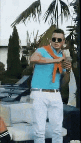 A Man In A Blue Polo Shirt And White Pants Is Standing In Front Of A Palm Tree Holding A Drink . GIF
