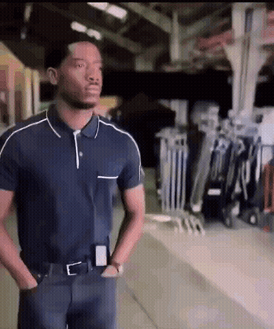 A Man In A Blue Polo Shirt Is Standing In A Warehouse With His Hands In His Pockets GIF