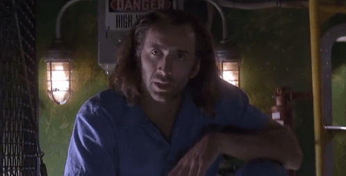 A Man In A Blue Scrub Is Standing In Front Of A Sign That Says Danger High Voltage GIF