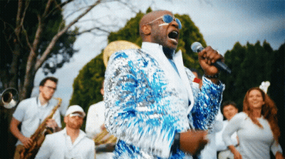 A Man In A Blue Sequined Jacket Sings Into A Microphone In Front Of A Band GIF