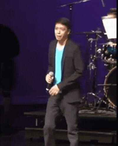 A Man In A Blue Shirt And A Black Jacket Is Singing Into A Microphone On A Stage GIF