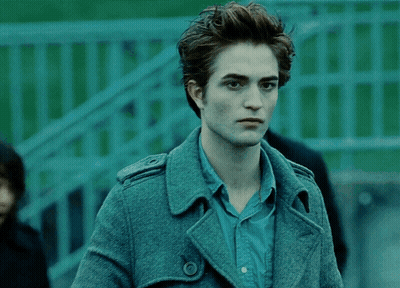 A Man In A Blue Shirt And A Grey Coat Stands In Front Of A Green Fence GIF
