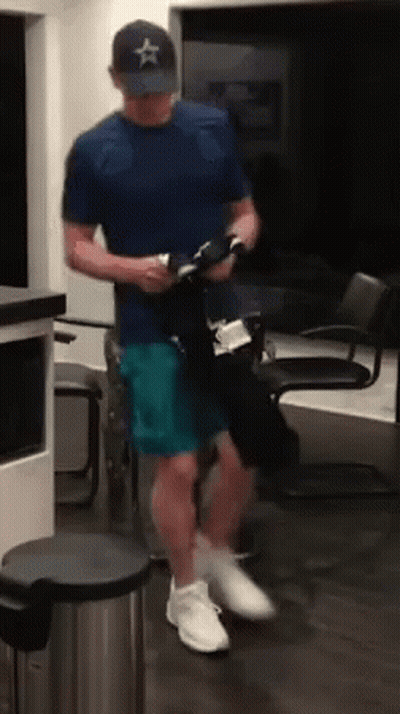 A Man In A Blue Shirt And A Hat Is Standing In A Room Holding A Bottle . GIF