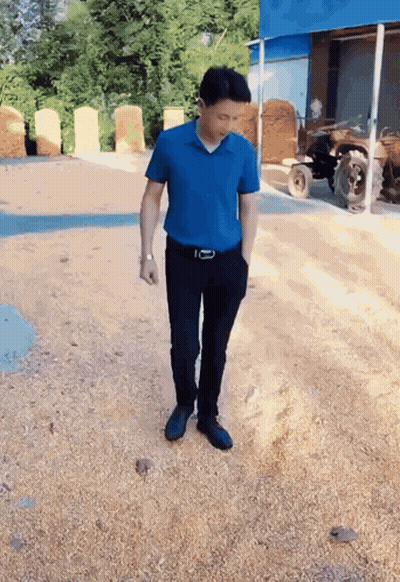 A Man In A Blue Shirt And Black Pants Is Standing On A Gravel Road GIF