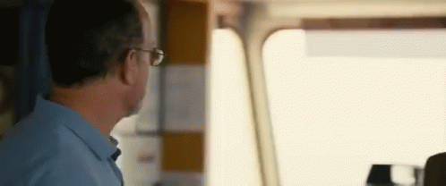 A Man In A Blue Shirt And Glasses Is Sitting In A Car Looking Out The Window . GIF