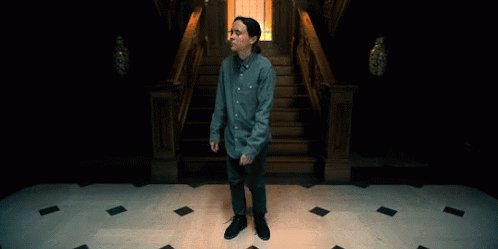 A Man In A Blue Shirt And Jeans Is Standing In Front Of A Set Of Stairs GIF
