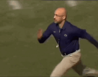 A Man In A Blue Shirt And Khaki Pants Is Running Across A Field . GIF