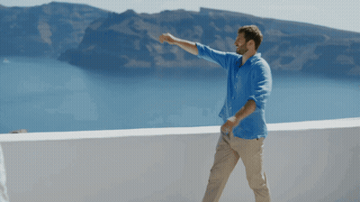A Man In A Blue Shirt And Khaki Pants Stands On A Balcony Overlooking The Ocean GIF
