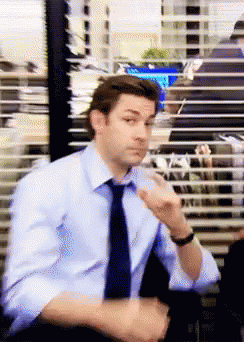 A Man In A Blue Shirt And Tie Is Sitting In Front Of A Window With Blinds On It GIF
