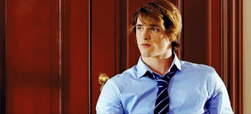 A Man In A Blue Shirt And Tie Is Sitting In Front Of A Wooden Wall . GIF