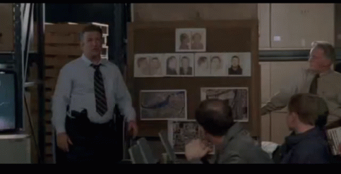 A Man In A Blue Shirt And Tie Stands In Front Of A Board With Pictures Of People On It GIF