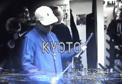 A Man In A Blue Shirt And White Hat Is Standing In A Room With The Word Kyoto On The Screen GIF