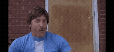 A Man In A Blue Shirt And White Shirt Is Sitting In Front Of A Brick Building . GIF