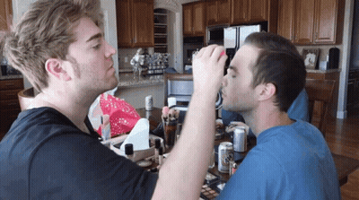 A Man In A Blue Shirt Is Applying Makeup To Another Man In A Kitchen GIF