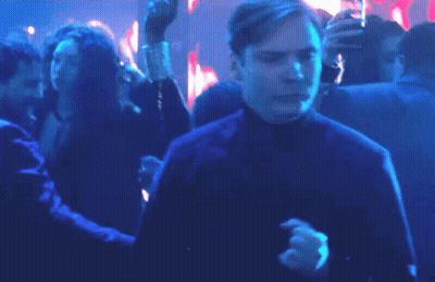 A Man In A Blue Shirt Is Dancing In A Crowd Of People In A Dark Room . GIF