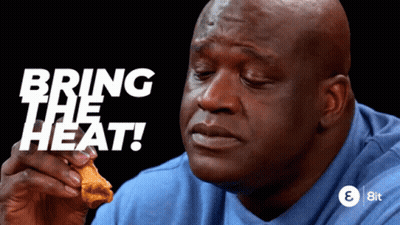 A Man In A Blue Shirt Is Eating A Chicken Wing With The Words " Bring The Heat " Behind Him GIF