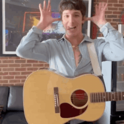 A Man In A Blue Shirt Is Holding A Guitar In Front Of A Brick Wall GIF