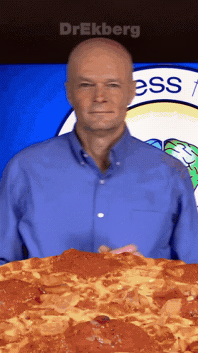 A Man In A Blue Shirt Is Holding A Large Pepperoni Pizza In Front Of A Sign That Says Ess GIF