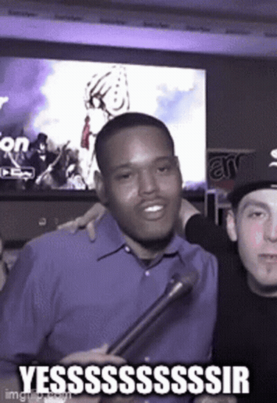 A Man In A Blue Shirt Is Holding A Microphone And Smiling While Another Man Looks On . GIF