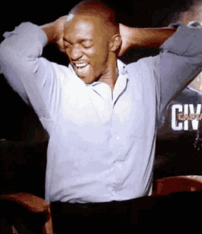 A Man In A Blue Shirt Is Laughing With His Hands Behind His Head In Front Of A Sign That Says City GIF