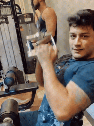 A Man In A Blue Shirt Is Lifting A Dumbbell In A Gym . GIF