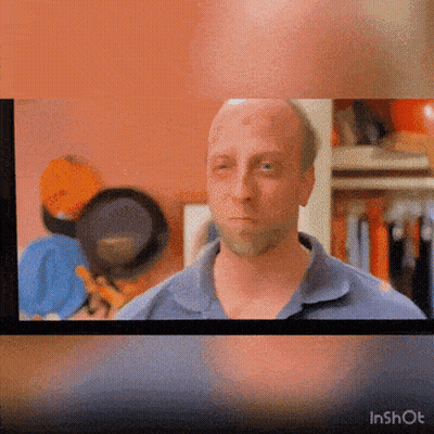 A Man In A Blue Shirt Is Looking At The Camera With A Pan Behind Him GIF