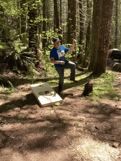 A Man In A Blue Shirt Is Playing A Game Of Cornhole In The Forest GIF