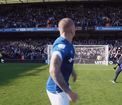 A Man In A Blue Shirt Is Running On A Soccer Field In Front Of A Sign That Says Umbro GIF