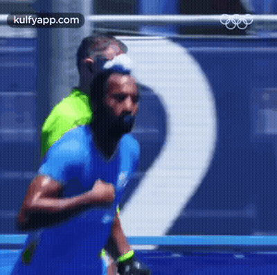 A Man In A Blue Shirt Is Running On A Track With A Man In A Green Shirt Behind Him . GIF