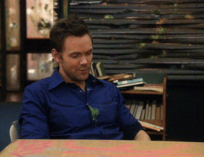 A Man In A Blue Shirt Is Sitting At A Table In Front Of A Stack Of Books GIF