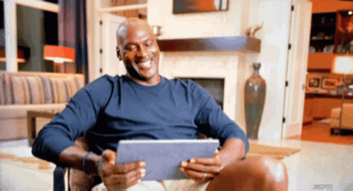 A Man In A Blue Shirt Is Sitting In A Chair Holding A Tablet And Smiling GIF