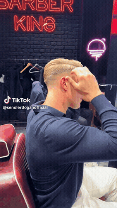 A Man In A Blue Shirt Is Sitting In Front Of A Neon Sign That Says Barber King GIF