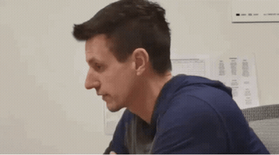 A Man In A Blue Shirt Is Sitting In Front Of A White Wall With Papers On It GIF