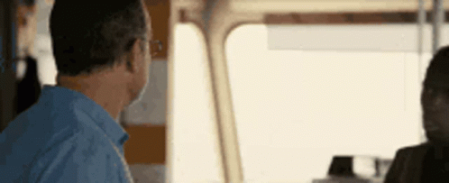 A Man In A Blue Shirt Is Sitting In Front Of A Window On A Boat Talking To Another Man . GIF