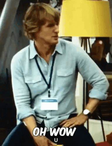 A Man In A Blue Shirt Is Sitting In Front Of A Yellow Lamp And Says Oh Wow GIF