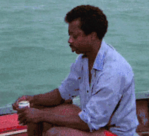 A Man In A Blue Shirt Is Sitting On A Boat Looking At The Water GIF