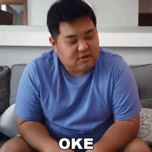 A Man In A Blue Shirt Is Sitting On A Couch And The Word Oke Is On His Chest GIF