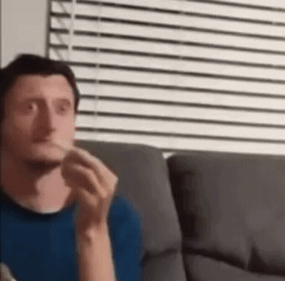 A Man In A Blue Shirt Is Sitting On A Couch Eating A Sandwich . GIF
