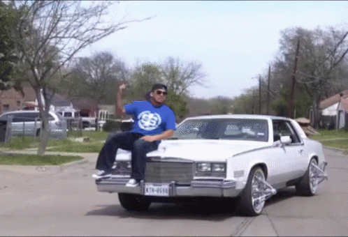A Man In A Blue Shirt Is Sitting On The Hood Of A White Car With A License Plate That Says K78-6030 GIF